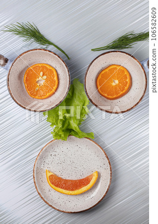 Ceramic dishes and an orange slice are folded in the form of a human face. 106582609