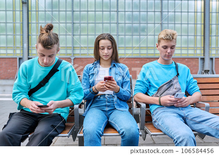Group of teenage friends using smartphones sitting on chairs on train station platform Group of teenage friends using smartphones sitting on chairs on train station platform 106584456
