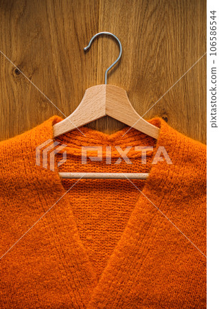 Autumn fall minimalist wardrobe with orange warm knitted sweater on wooden clothes hanger. Autumn capsule, warm fashion trends and stylish warm background, knitwear, copy space. Autumn fall minimalist wardrobe with orange warm knitted sweater on wooden clothes hanger. Autumn capsule, warm fashion trends and stylish warm background, knitwear, copy space. 106586544