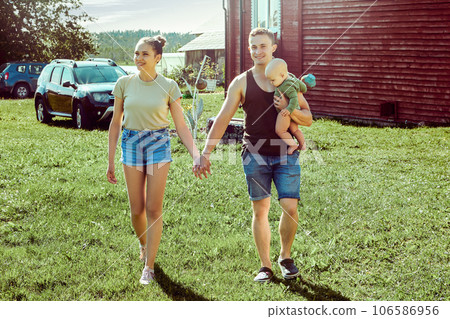 Young family couple with baby walks near country house holding hands on sunny summer day. 106586956