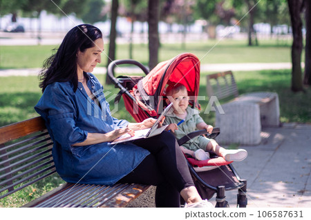 Brunette woman working online via laptop and cute baby boy sitting calmly in buggy in summer park single mother searching data on internet with infant child in garden 106587631