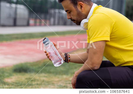 Upset man with wireless headphones looking at fresh water bottle sitting after workout at sports arena sportsman in color t-shirt holding drink during training break Upset man with wireless headphones looking at fresh water bottle sitting after workout at sports arena sportsman in color t-shirt holding drink during training break 106587641