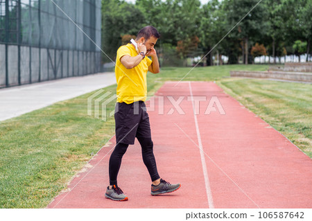 Man in tracksuit putting on cordless earphones walking along track at urban sports arena thoughtful athlete in yellow t-shirt training with music at outdoor ground Man in tracksuit putting on cordless earphones walking along track at urban sports arena thoughtful athlete in yellow t-shirt training with music at outdoor ground 106587642