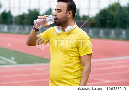 Man in yellow sportive t-shirt drinking from open water bottle and resting after training on running track at outdoor sports ground bearded sportsman refreshing with drink Man in yellow sportive t-shirt drinking from open water bottle and resting after training on running track at outdoor sports ground bearded sportsman refreshing with drink 106587643