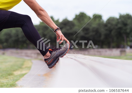 Man wearing black leggings touching sports shoe toe cap leaning on bench and doing exercises in spring urban park flexible man training leg muscles outdoors 106587644
