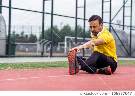 Athlete with headphones touching foot toes sitting on running track at outdoor modern stadium in city park sportsman stretching leg muscles on urban sports arena Athlete with headphones touching foot toes sitting on running track at outdoor modern stadium in city park sportsman stretching leg muscles on urban sports arena 106587647