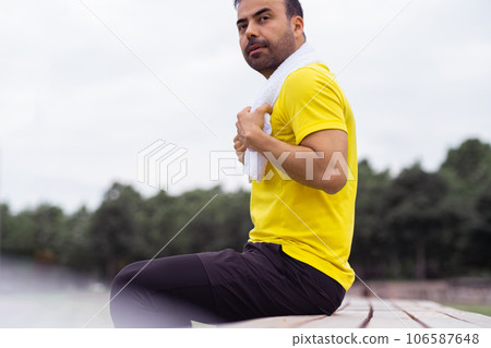 Handsome man in tracksuit with white towel on neck sitting on wooden bench in green city park sportsman resting during routine training on outdoor sports ground 106587648