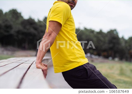 working on arm muscles, man in yellow t-shirt performs backward push-ups, using a bench in the serene green city park as his exercise platform on the dynamic sports arena. working on arm muscles, man in yellow t-shirt performs backward push-ups, using a bench in the serene green city park as his exercise platform on the dynamic sports arena. 106587649