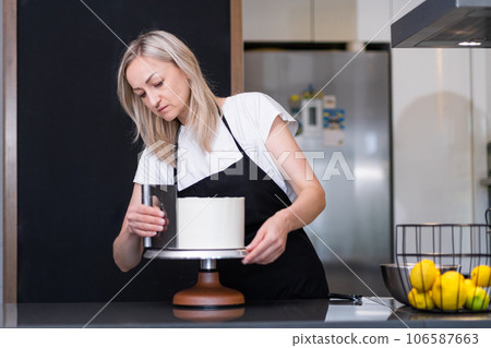 Focused female blogger in black apron using pastry scraper to process white mastic on cake blonde woman standing at kitchen table showing followers process of confectionery preparation Focused female blogger in black apron using pastry scraper to process white mastic on cake blonde woman standing at kitchen table showing followers process of confectionery preparation 106587663