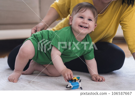 Happy baby boy smiles playing with vehicle toy sitting on soft white rug near loving mother woman enjoying entertaining chubby little son relaxing at home Happy baby boy smiles playing with vehicle toy sitting on soft white rug near loving mother woman enjoying entertaining chubby little son relaxing at home 106587667