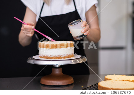 Professional confectioner using pink brush to smear cake layers with cream in glass jar woman in black apron cooking custom confectionery for anniversary celebration standing at table Professional confectioner using pink brush to smear cake layers with cream in glass jar woman in black apron cooking custom confectionery for anniversary celebration standing at table 106587670