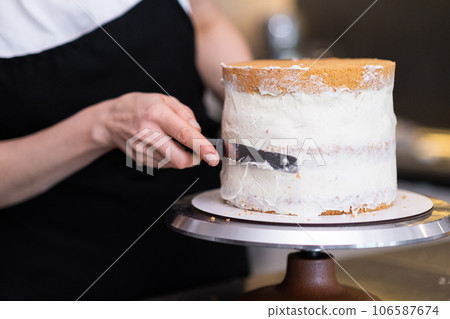 Skilled confectioner smearing sugar free white cream around baked cake with knife on blurred background woman in black apron creating delicious custom pastry for events closeup Skilled confectioner smearing sugar free white cream around baked cake with knife on blurred background woman in black apron creating delicious custom pastry for events closeup 106587674