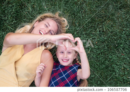 Smiling mother and little daughter lie on green grass on sunny summer day young woman and girl make heart symbol with hands with happy expressions on faces upper view 106587706