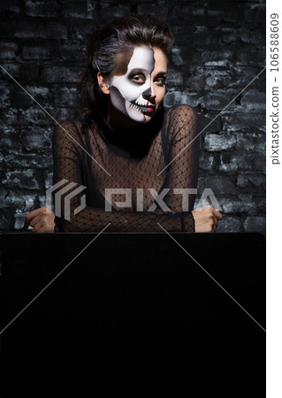 Young female wearing scary skeleton makeup leaning to copy space. Front view of brunette woman with skull Halloween facepaint looking to camera with light smile. Concept of Halloween, face art. Young female wearing scary skeleton makeup leaning to copy space. Front view of brunette woman with skull Halloween facepaint looking to camera with light smile. Concept of Halloween, face art. 106588609