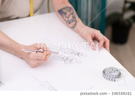 Lady designer draws clothes model on paper at table in fashion atelier tailor makes pencil sketch of apparel for women in sewing studio closeup dressmaking industry Lady designer draws clothes model on paper at table in fashion atelier tailor makes pencil sketch of apparel for women in sewing studio closeup dressmaking industry 106589281