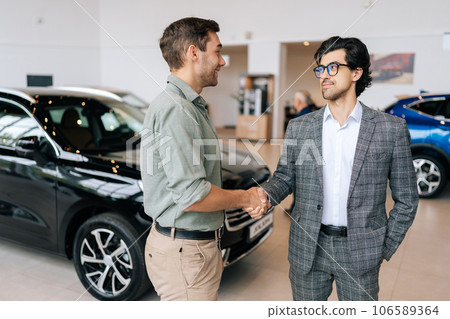 Portrait of confident car dealer in business suit welcoming young man customer in showroom. Professional salesman shaking client hand in auto dealership. Concept of buying new car. Portrait of confident car dealer in business suit welcoming young man customer in showroom. Professional salesman shaking client hand in auto dealership. Concept of buying new car. 106589364