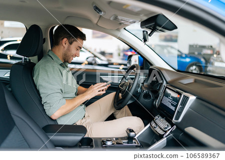 Side view of happy handsome young man sitting behind wheel of luxury car and using mobile phone for surfing internet social media e-mail. Smiling bearded businessman using smartphone app at showroom. 106589367