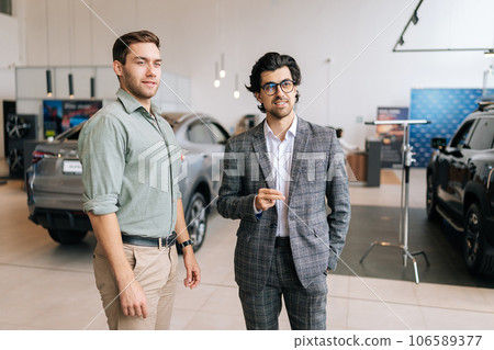Portrait of handsome male client talking to positive car dealer wearing business suit in dealership discussing automobiles looking at luxurious new model. Concept of choosing and buying new auto Portrait of handsome male client talking to positive car dealer wearing business suit in dealership discussing automobiles looking at luxurious new model. Concept of choosing and buying new auto 106589377