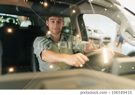 Portrait through windshield of happy customer male client putting hand on steering wheel, smiling looking at camera while trying new automobile. Happy young man client doing drive test of new car. Portrait through windshield of happy customer male client putting hand on steering wheel, smiling looking at camera while trying new automobile. Happy young man client doing drive test of new car. 106589418