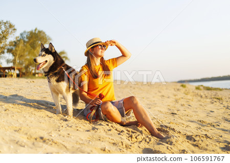 Woman in hat and sunglasses sits with her dog on the beach and enjoying sunset Woman in hat and sunglasses sits with her dog on the beach and enjoying sunset 106591767
