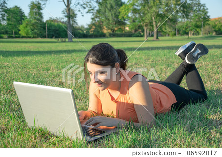 Caucasian Woman lying on park wearing light fitness clothing using laptop happy Caucasian Woman lying on park wearing light fitness clothing using laptop happy 106592017