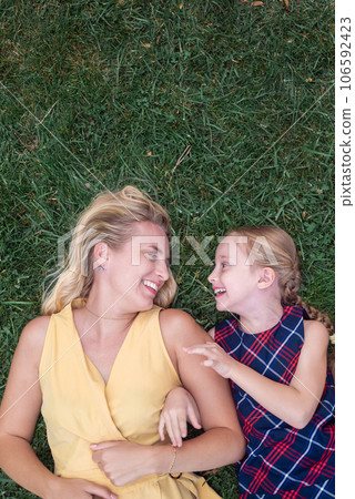 From above, a blonde mother and her daughter lie on lush green grass in the park, sharing joyful laughter, creating a heartwarming scene.  106592423