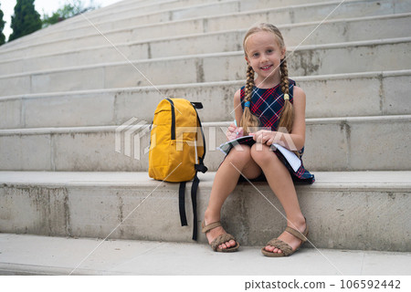 In the schoolyard, a studious young girl with her schoolbag diligently works on tasks, writing in her notebook on stone steps.  106592442