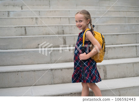 Cute girl with long braids smiles looking at camera schoolgirl in colorful dress poses holding strap of yellow backpack child goes to school climbing concrete stairs 106592443