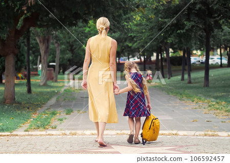 A lively schoolgirl, her yellow backpack bouncing, playfully hops while walking with her mother across the park on their way to school.  106592457