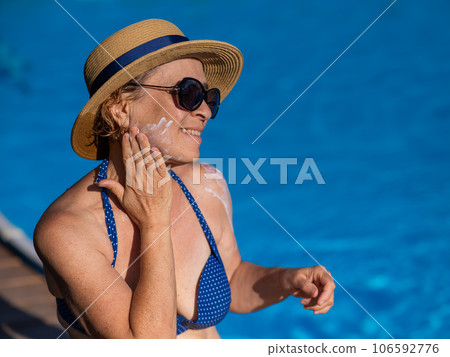 Portrait of an old woman in a straw hat, sunglasses and a swimsuit applying sunscreen to her face while relaxing by the pool.  106592776
