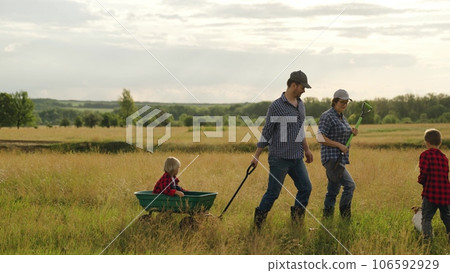 Farmer parents ride little boy in cart and meet elder son in large field at sunset. Farmer family with children goes to work along meadow at farmland. Farmers with little kids and tools walk in field 106592929