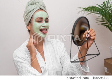 woman in bathrobe joyfully admires her reflection in mirror with facial cosmetic mask showing tongue to mirror  106593120