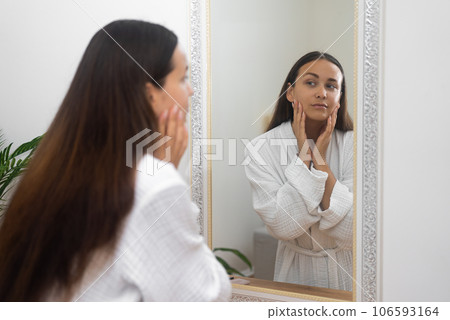 Attractive brunette woman in gown looking in mirror in bathroom lady checking facial skin condition standing by dressing table at home woman with healthy skin near mirror Attractive brunette woman in gown looking in mirror in bathroom lady checking facial skin condition standing by dressing table at home woman with healthy skin near mirror 106593164