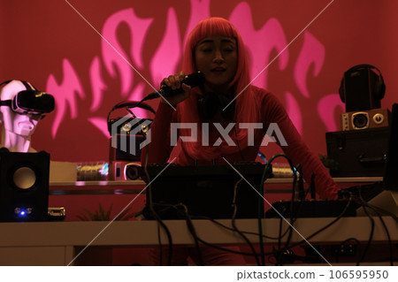 Cheeful artist with pink hair standing at dj table playing electronic music with turntables, enjoying to perform at night in club. Asian performer having fun with fans during techno concert 106595950