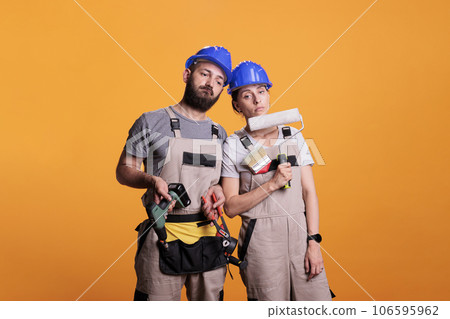 Exhausted team of contractors working with construction tools, falling asleep after renovating project. Man and woman builders holding power drill, painting brush or roller and pair of pliers. Exhausted team of contractors working with construction tools, falling asleep after renovating project. Man and woman builders holding power drill, painting brush or roller and pair of pliers. 106595962