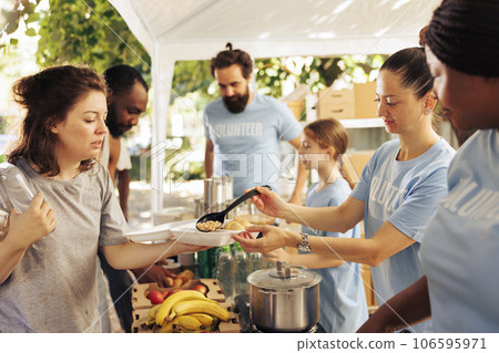 Multicultural individuals volunteering at an outdoor food bank serving warm meals, non-perishable items, and support for homeless people. Humanitarian aid provided to the poor and needy. 106595971