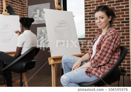 Happy young woman sitting at easel in classroom and smiling at camera, enjoying fun drawing class with friends, learning sketching techniques at group art class. Enjoyable creative hobby 106595976