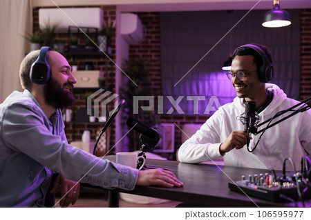 Online show host recording podcast with guest, using audio recording equipment in home studio living room. Happy men laughing together during broadcasting session for internet streaming service 106595977
