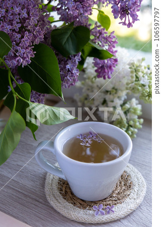 Tasty black tea in white cup on windowsill with aromatic lilac flowers. Spring composition Cup of lilac tea drinking recipe flowering branches of purple lilac. Still life for copy space greeting card Tasty black tea in white cup on windowsill with aromatic lilac flowers. Spring composition Cup of lilac tea drinking recipe flowering branches of purple lilac. Still life for copy space greeting card 106597797
