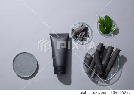 Black tube without label decorated with some petri dishes containing activated charcoal sticks, clay mask and green tea leaves. Empty label for cosmetic product mockup 106605252
