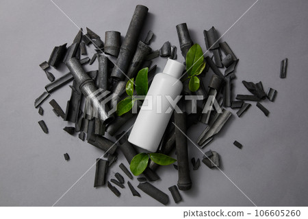 Blank label bottle placed on a pile of activated charcoal sticks. Few green tea leaves decorated on gray background. Mockup of skin care cosmetic bottle of beauty facial 106605260