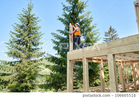 Back view of man wearing yellow vest and helmet, standing high, building wooden house in forest. Builder, craftsman, engineer, holding, measuring wooden gilder. Concept of forest. Back view of man wearing yellow vest and helmet, standing high, building wooden house in forest. Builder, craftsman, engineer, holding, measuring wooden gilder. Concept of forest. 106605518