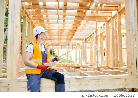 Side view of man wearing uniform sitting on wooden decking, holding sheet of paper, constructing. Young architect, engineer, builder planning project. Concept of building. Side view of man wearing uniform sitting on wooden decking, holding sheet of paper, constructing. Young architect, engineer, builder planning project. Concept of building. 106605538