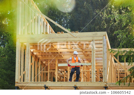 Front view of man, builder, worker building wooden house in forest, in sunny weather, in summer. Male wearing helmet and uniform holding, putting gilder. Concept of building. 106605542
