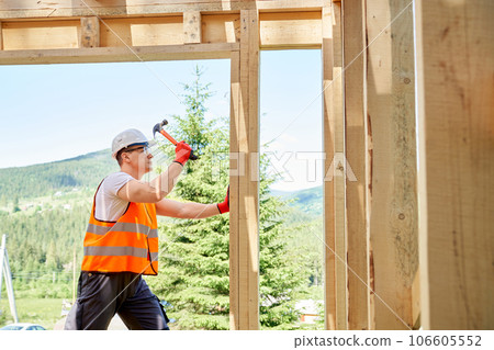 Side view of professional worker, builder standing, holding hammer and wooden gilder, beating. Man wearing vest and helmet, building wooden house in forest. Concept of building. Side view of professional worker, builder standing, holding hammer and wooden gilder, beating. Man wearing vest and helmet, building wooden house in forest. Concept of building. 106605552