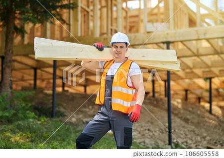 Front view of man, worker wearing uniform and helmet, standing, holding wooden boards, gilders. Builder constructing wooden house, looking at camera. Concept of building. Front view of man, worker wearing uniform and helmet, standing, holding wooden boards, gilders. Builder constructing wooden house, looking at camera. Concept of building. 106605558
