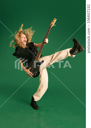 Full-length image of young girl in stylish clothes emotionally playing electric guitar against green studio background Full-length image of young girl in stylish clothes emotionally playing electric guitar against green studio background 106607282