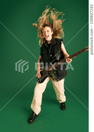 Full-length image of young girl in stylish clothes emotionally playing electric guitar against green studio background Full-length image of young girl in stylish clothes emotionally playing electric guitar against green studio background 106607283
