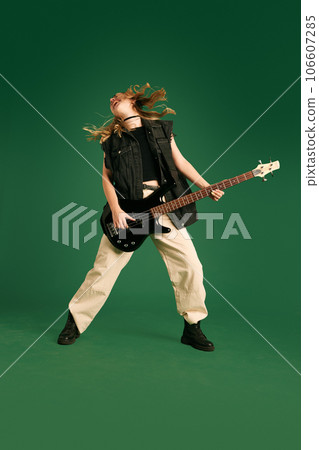 Full-length image of young girl in stylish clothes emotionally playing electric guitar against green studio background Full-length image of young girl in stylish clothes emotionally playing electric guitar against green studio background 106607285