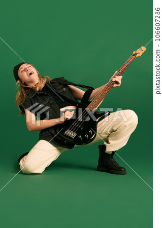 Emotional young girl in stylish clothes standing on one knee and playing electric guitar against green studio background Emotional young girl in stylish clothes standing on one knee and playing electric guitar against green studio background 106607286